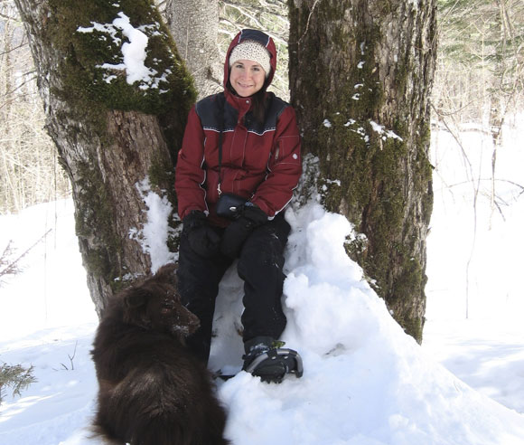 Photo d'Édith Bégin en hiver, entre deux arbres, avec un chien aux longs poils bruns à ses pieds